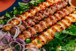 A wooden dining table topped with authentic Persian kabob platters, featuring skewers of ground beef koobideh and saffron chicken with charred tomatoes, fluffy basmati rice, flatbread, and fresh sides.